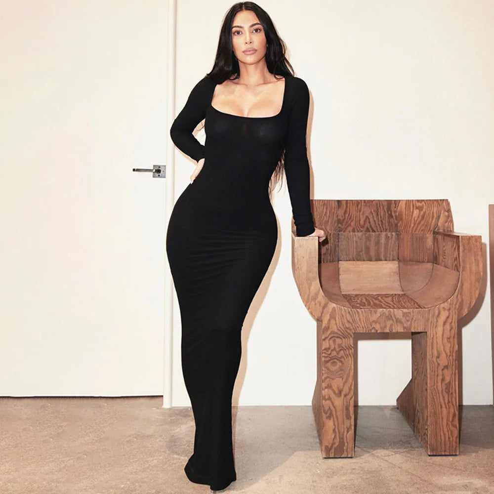 Woman in a black dress standing next to a wooden chair against a white wall.